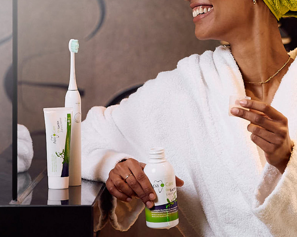A smiling woman in a white bathrobe and green towel headwrap holds a small white bottle with a green label in a bathroom setting. An electric toothbrush and a tube of toothpaste are also visible on a dark countertop beside a mirror.