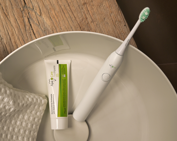 A white electric toothbrush with green bristles rests in a white ceramic bowl alongside a tube of toothpaste and a textured white towel, set on a wooden surface.