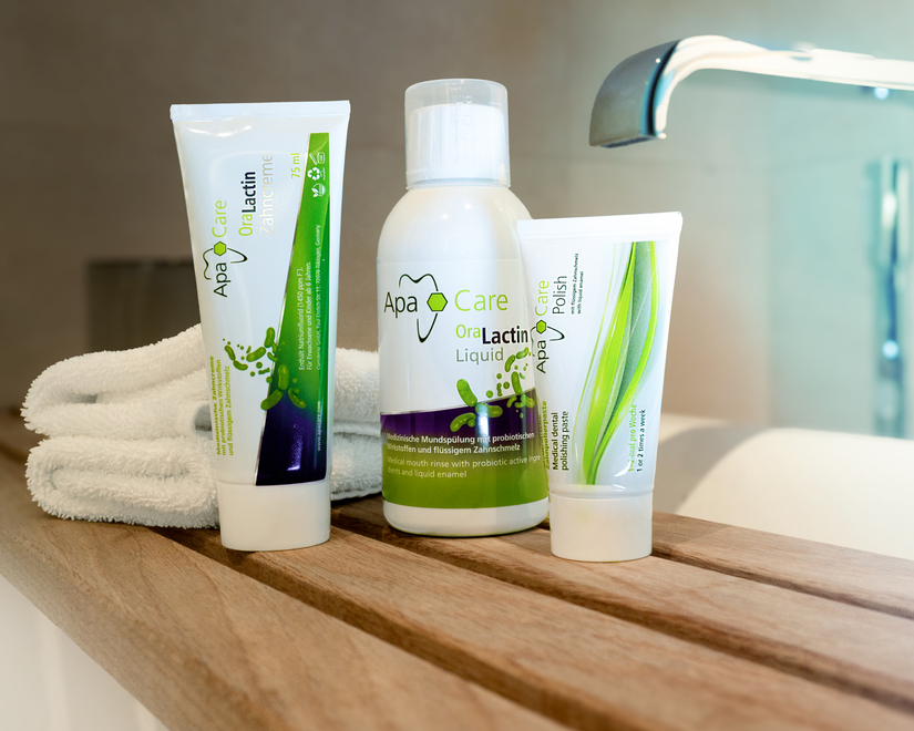 Three dental care products from Apa Care Organic Lactin are arranged on a wooden bathroom shelf next to a folded white towel and a sink faucet.